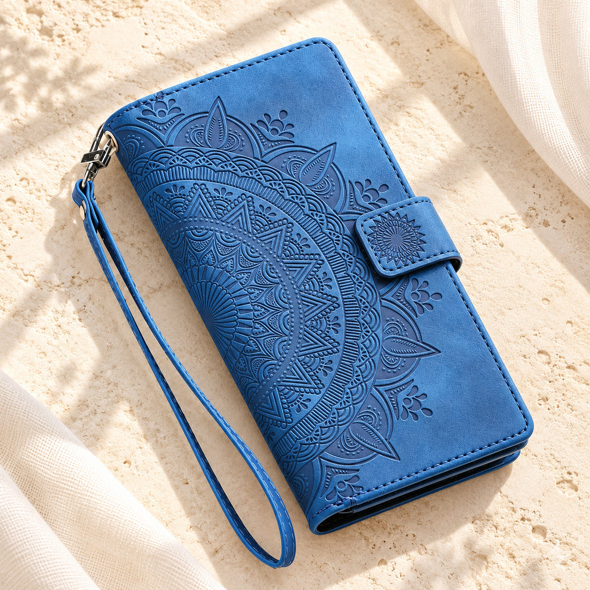 Lotus Mandala iPhone wallet case in Blue showing the closed embossed cover and matching wrist loop on limestone
