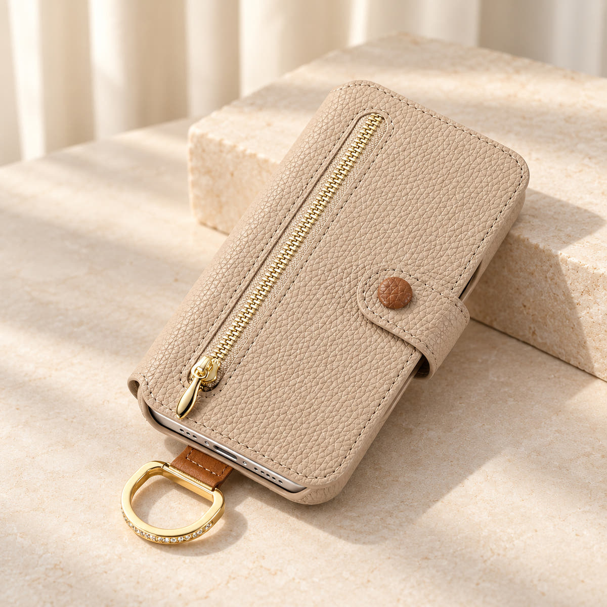 Beige textured iPhone wallet case closed on a cream surface, showing the front zip pocket, buttoned tab and attached gold-tone D-ring