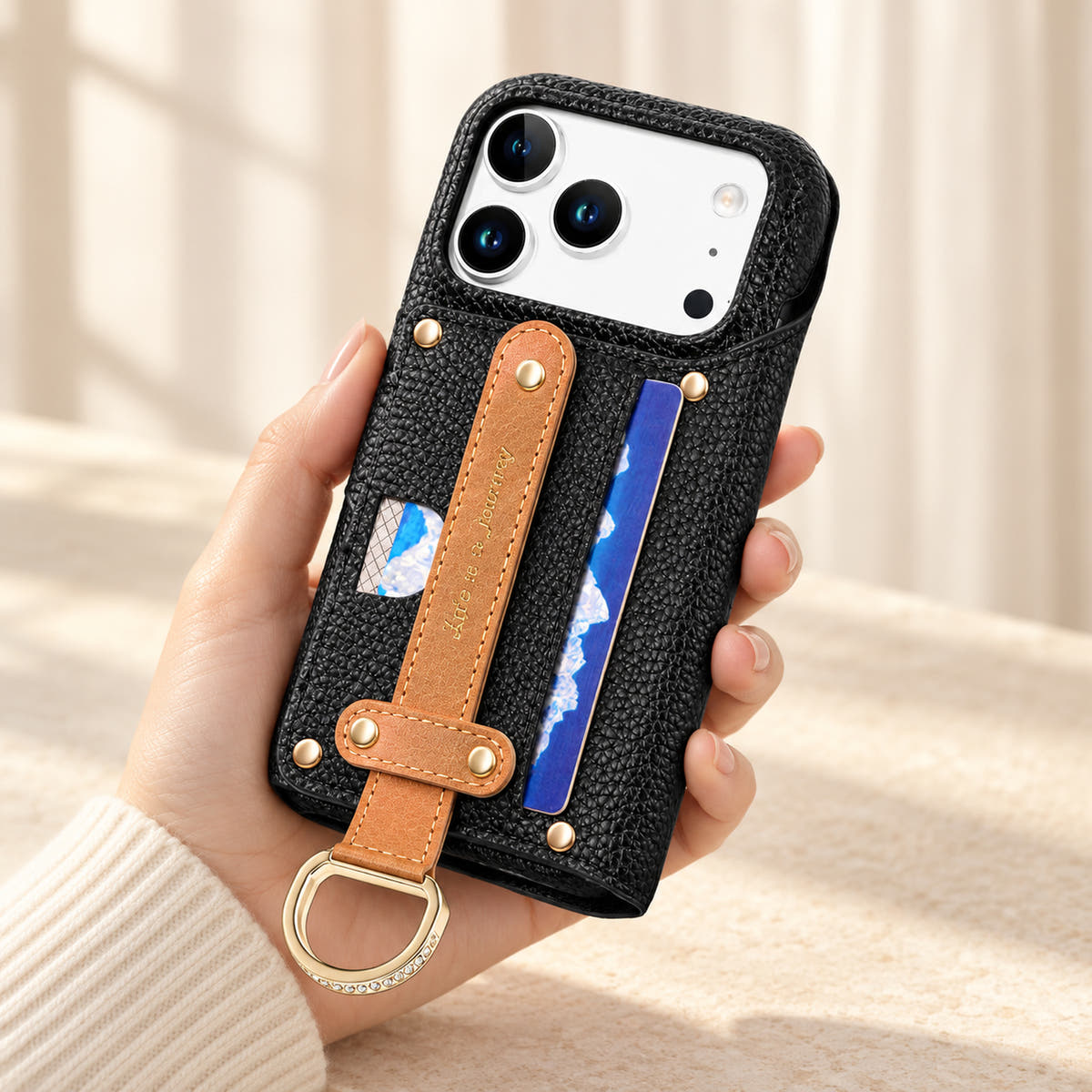 Black textured iPhone wallet case held in hand, showing the contrasting rear hand strap, card pocket, gold-tone D-ring and camera opening