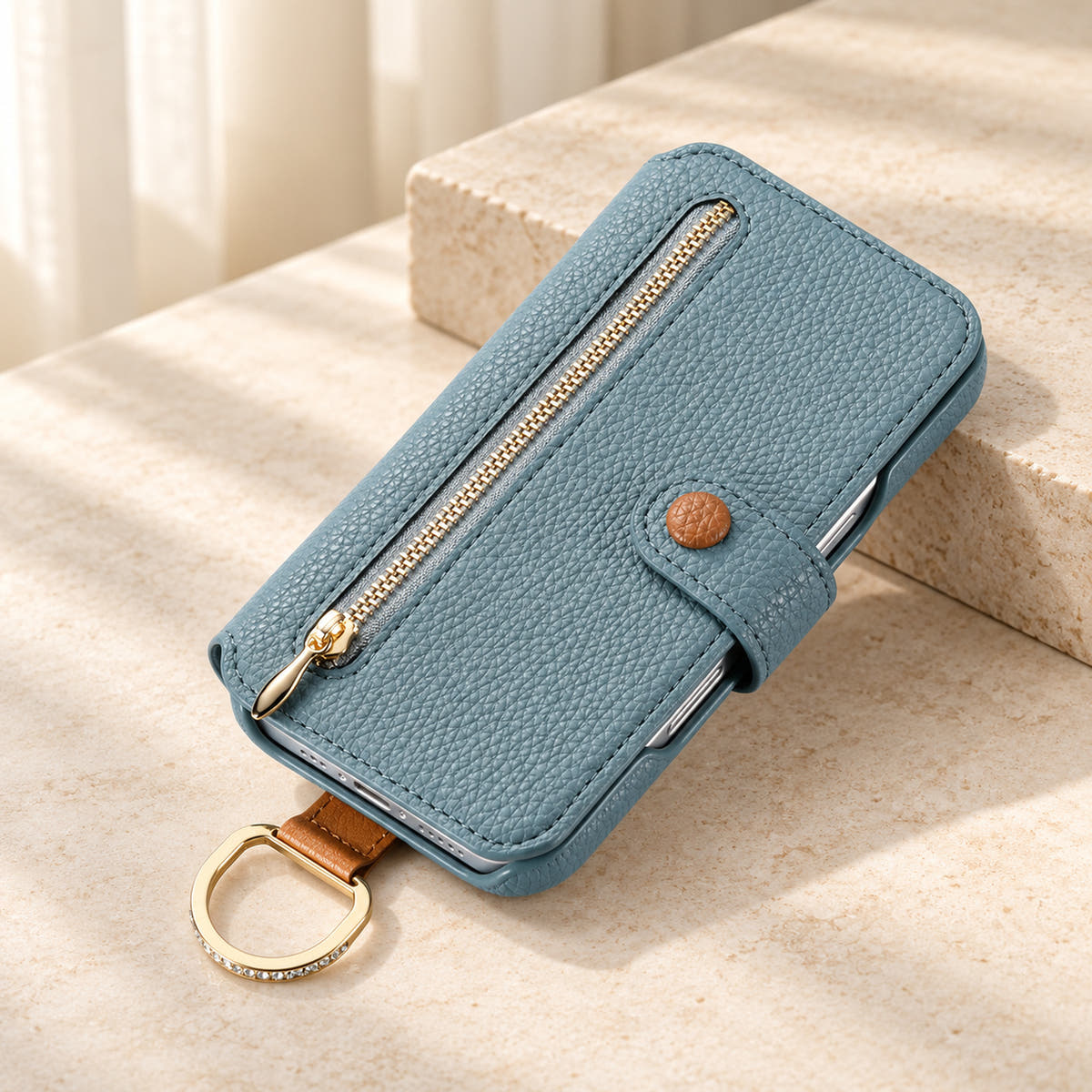 Blue textured iPhone wallet case closed on a cream surface, showing the front zip pocket, buttoned tab and attached gold-tone D-ring