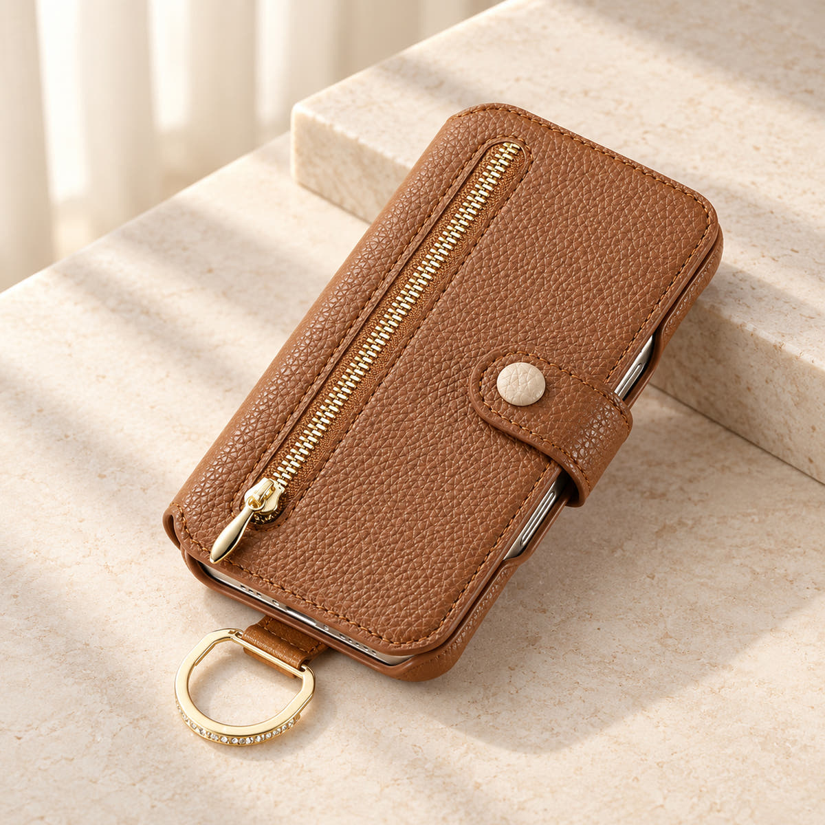 Brown textured iPhone wallet case closed on a cream surface, showing the front zip pocket, buttoned tab and attached gold-tone D-ring