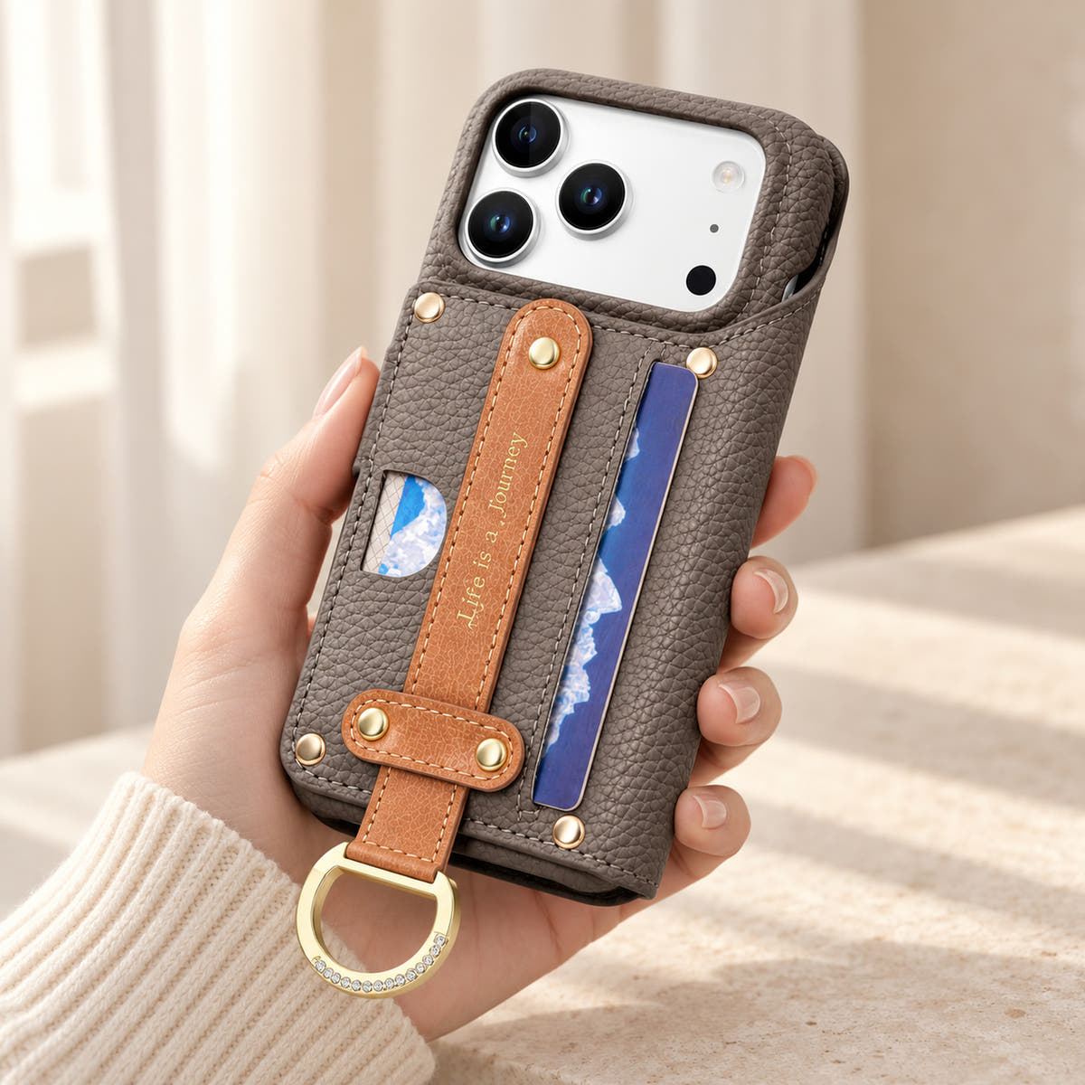 Gray textured iPhone wallet case held in hand, showing the contrasting rear hand strap, card pocket, gold-tone D-ring and camera opening