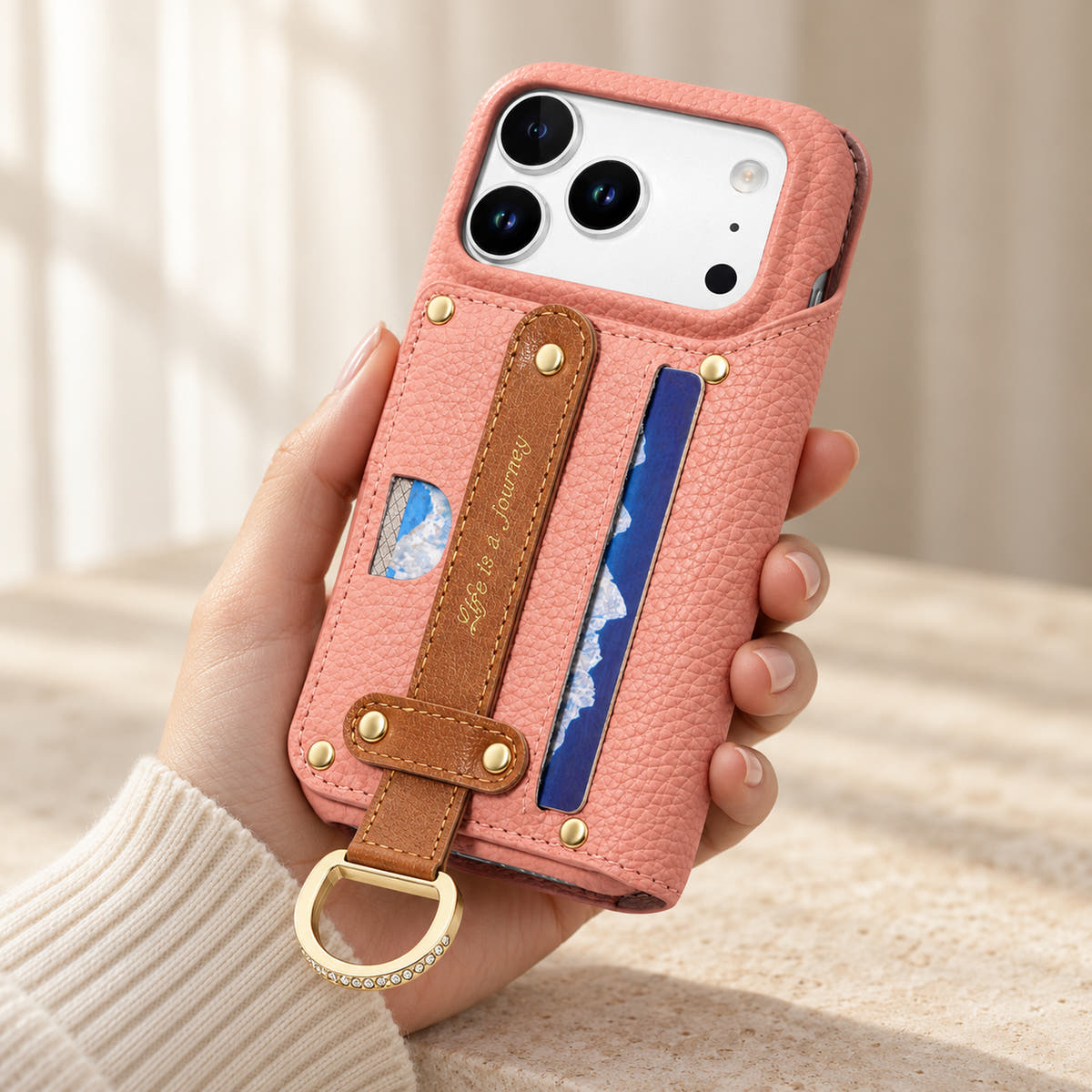 Pink textured iPhone wallet case held in hand, showing the contrasting rear hand strap, card pocket, gold-tone D-ring and camera opening