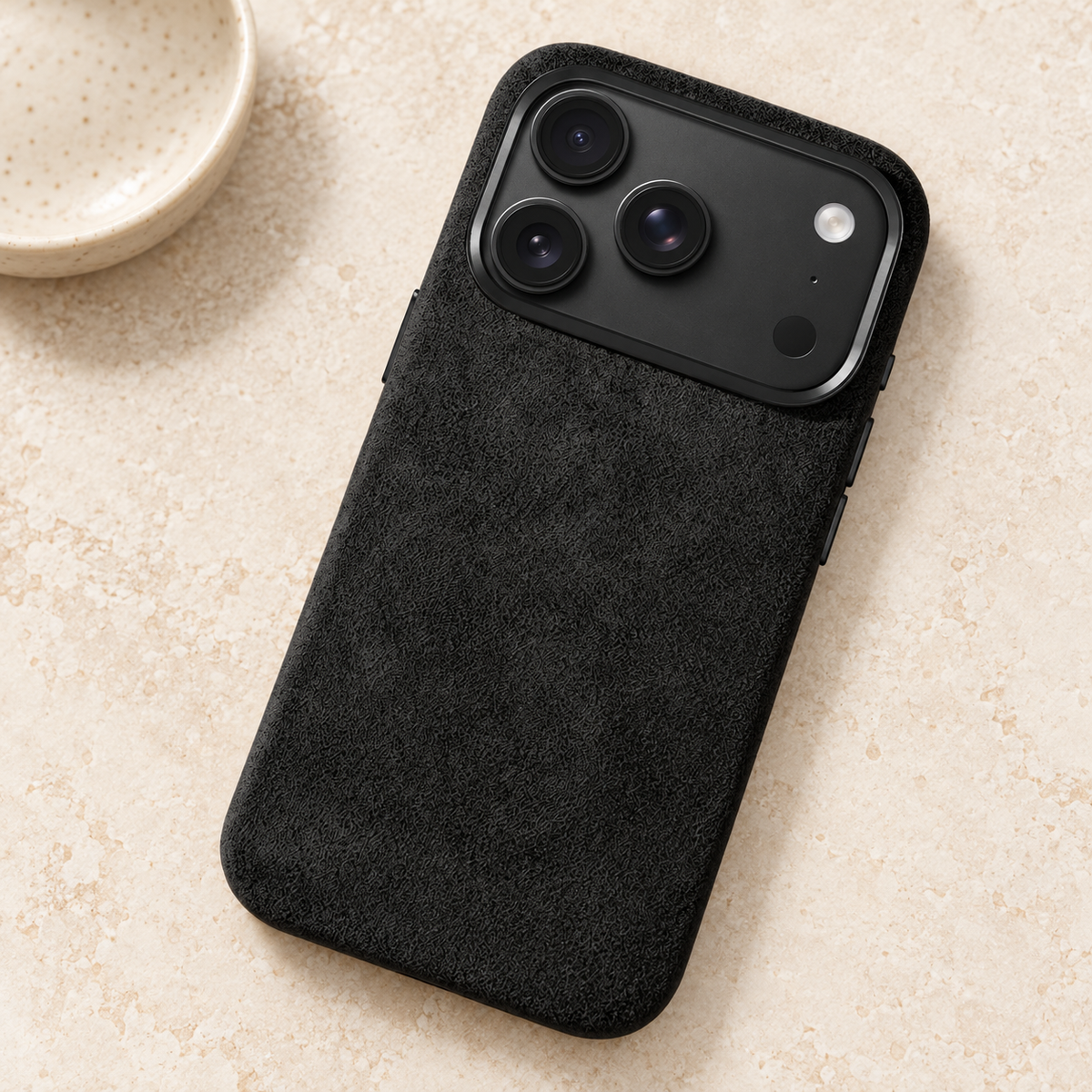 Black suede-look textured iPhone case resting on a warm cream stone tabletop.