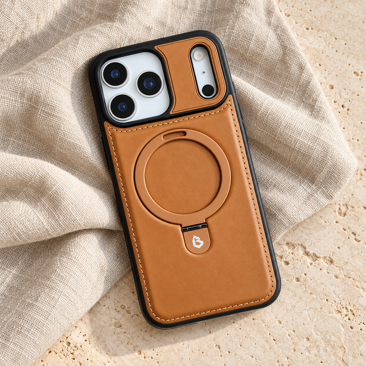 Tan stitched ring-stand iPhone case in a diagonal flatlay on oatmeal linen and cream stone.