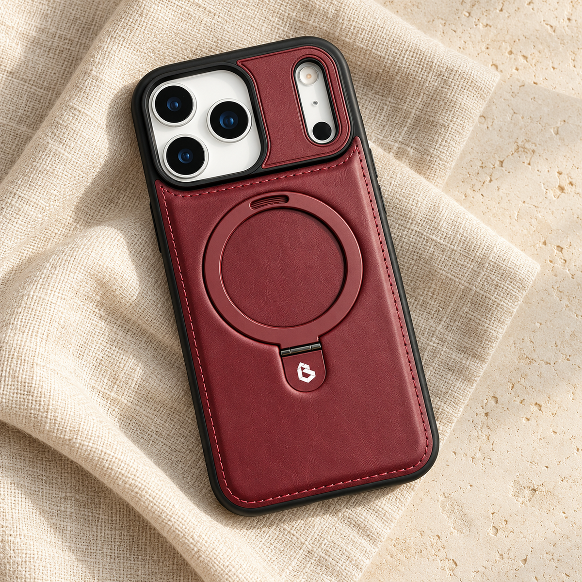 Burgundy stitched ring-stand iPhone case in a diagonal flatlay on oatmeal linen and cream stone.