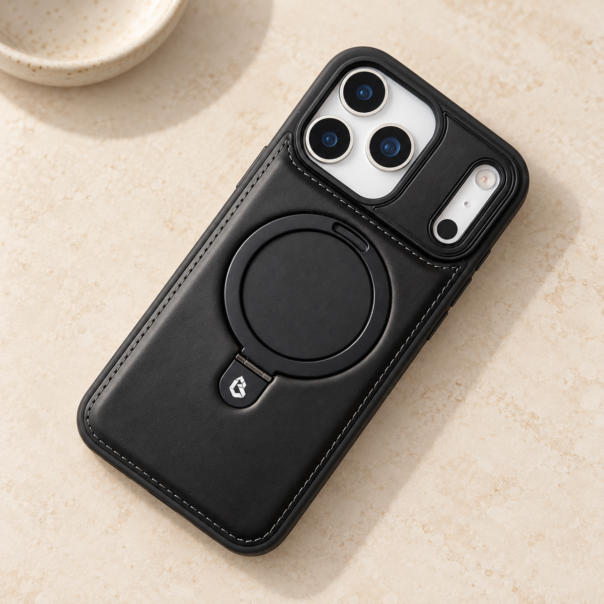 Black stitched ring-stand iPhone case resting on a warm cream stone tabletop.