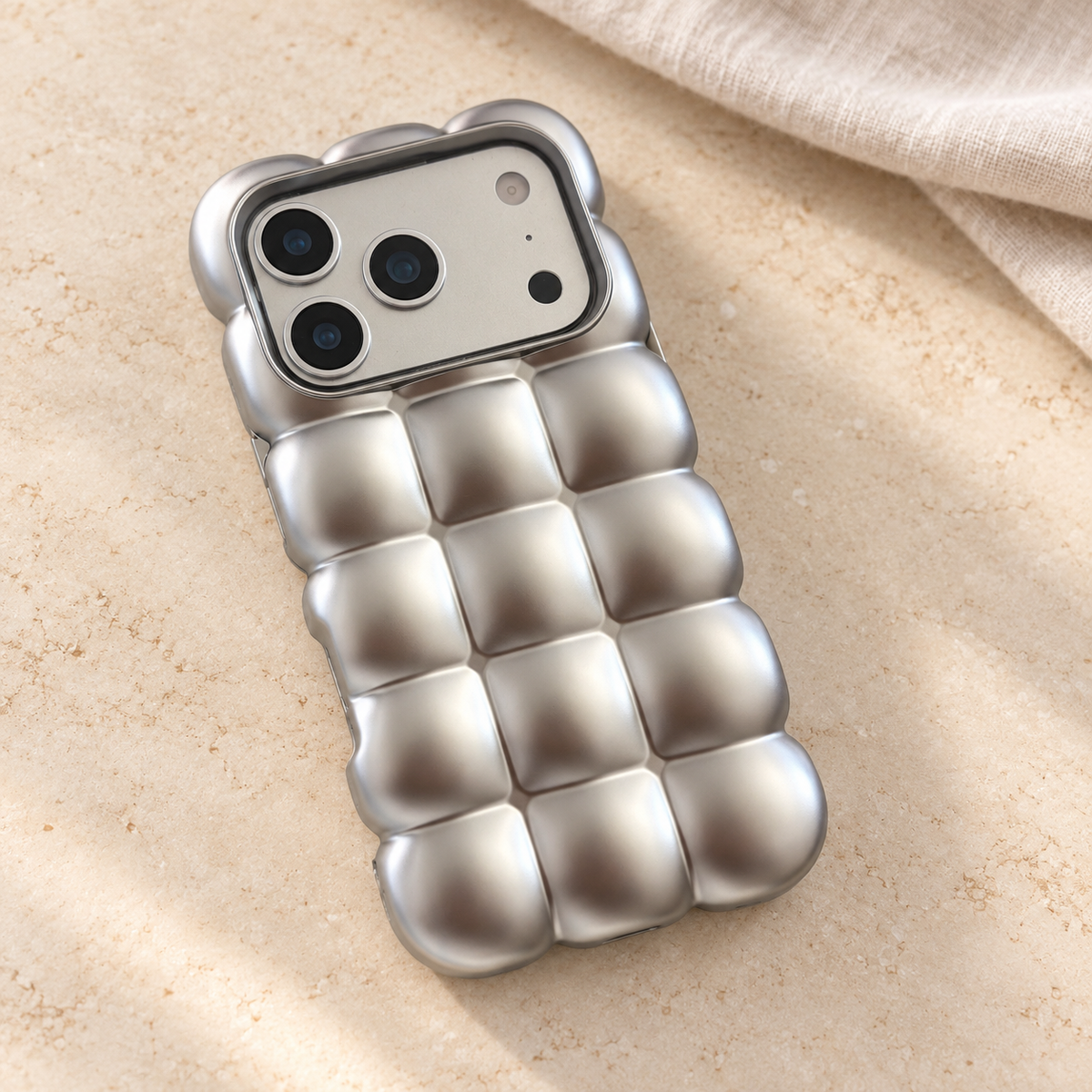 Satin Silver puffy grid iPhone case with rounded-square back and scalloped edges — diagonal tabletop view.