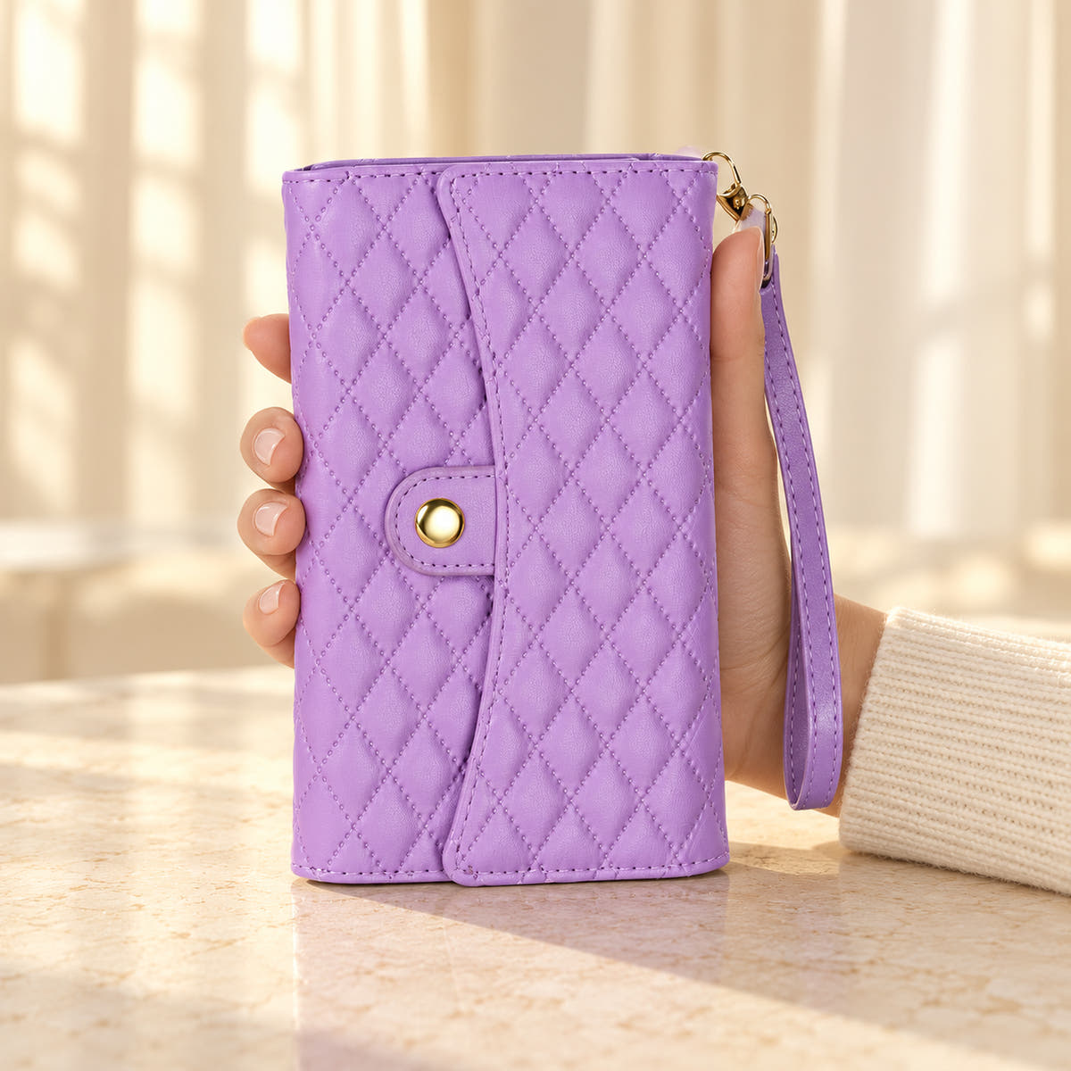 Quilted Zip Wallet iPhone case in Lavender held in a cream-sleeved hand, showing its diamond-quilted flap, gold-tone snap and matching wrist strap.