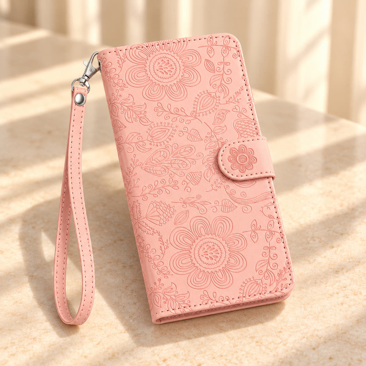 ScarletBloom Pink wallet phone case on a sunlit cream limestone tabletop, showing the embossed floral cover and matching wrist loop.