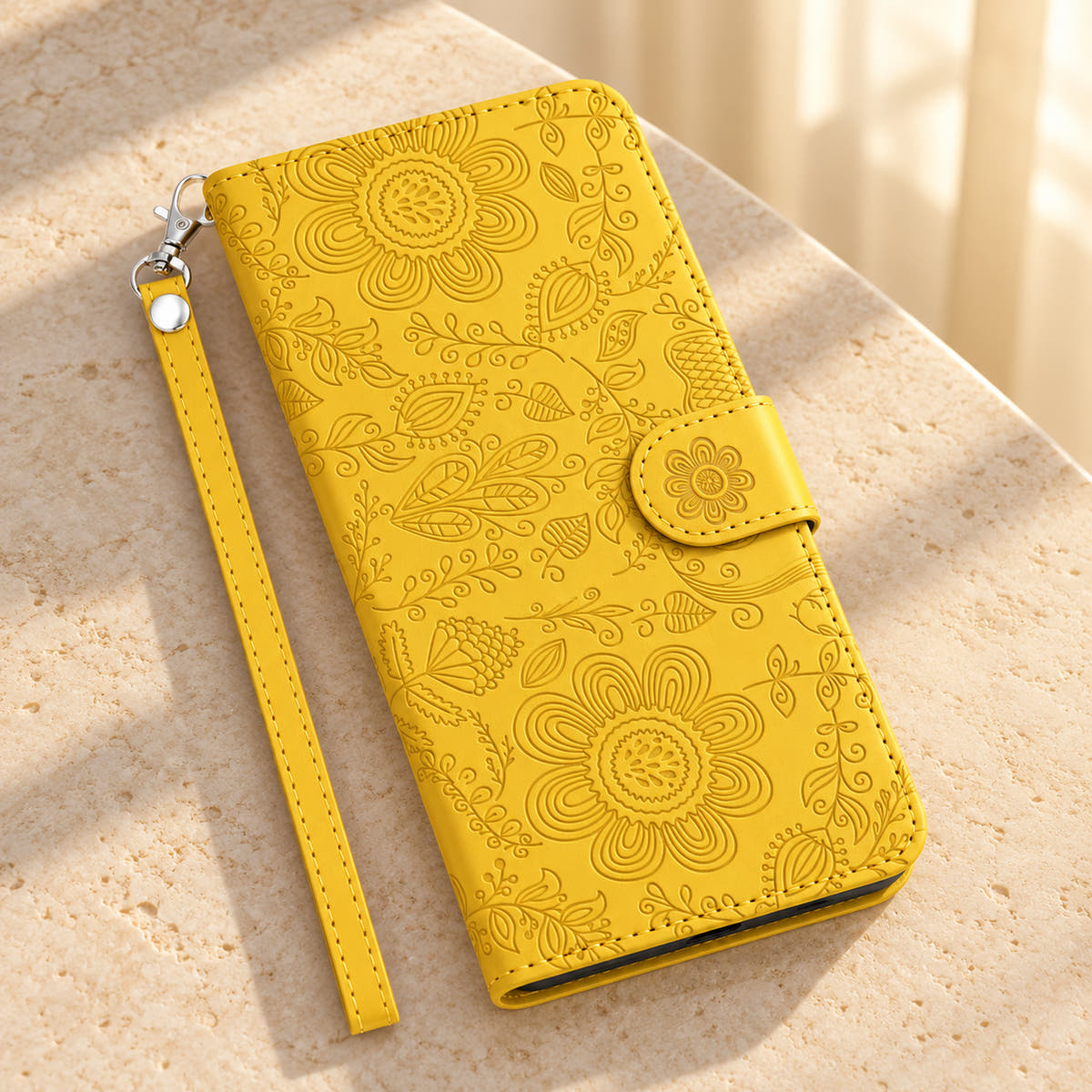 Yellow Scarlet Bloom wallet phone case on a sunlit cream stone tabletop with floral embossing, a flower-decorated closure tab and a matching wrist loop.