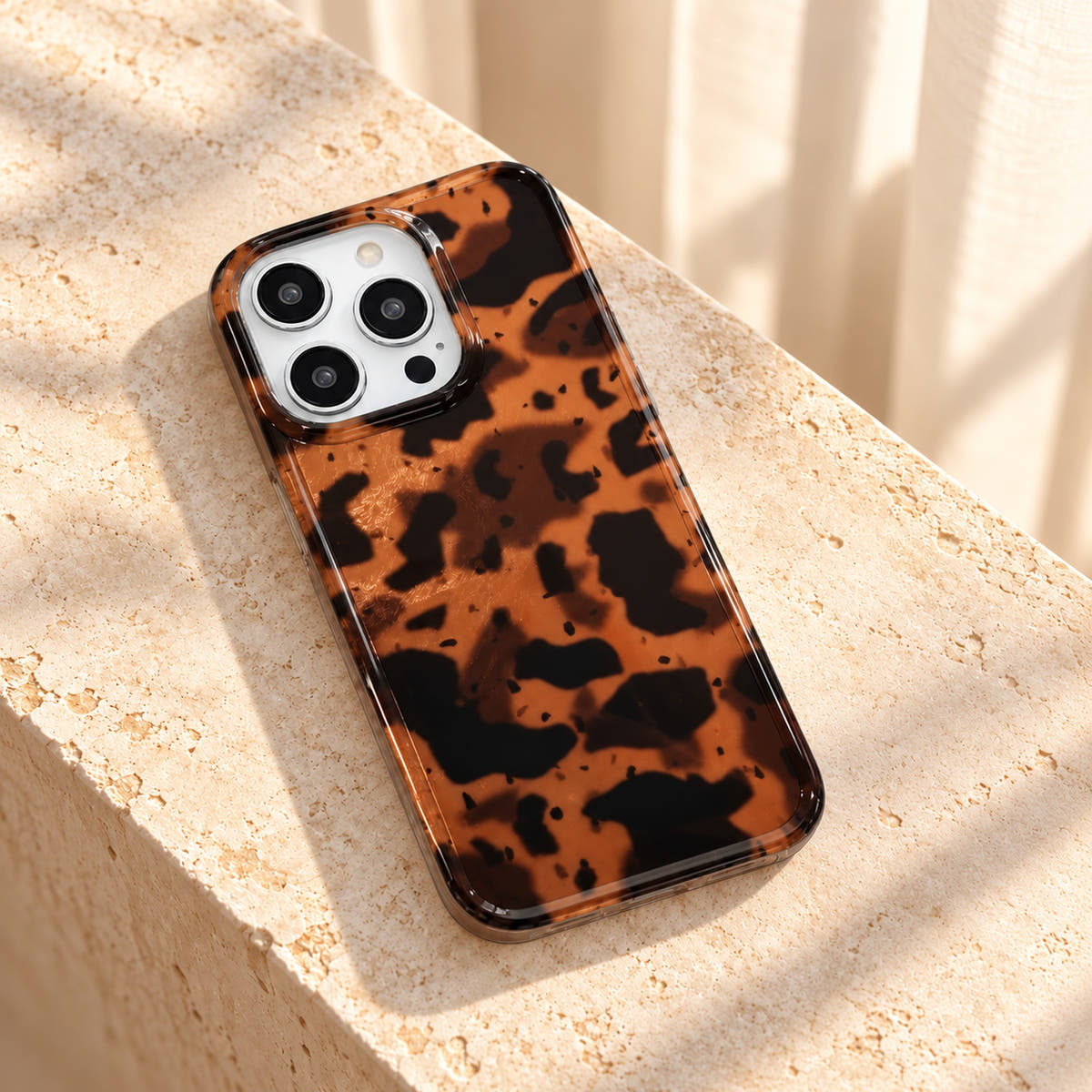 Sienna Luxe Brown tortoiseshell iPhone case resting diagonally on cream limestone with its glossy rear and clear edge visible.