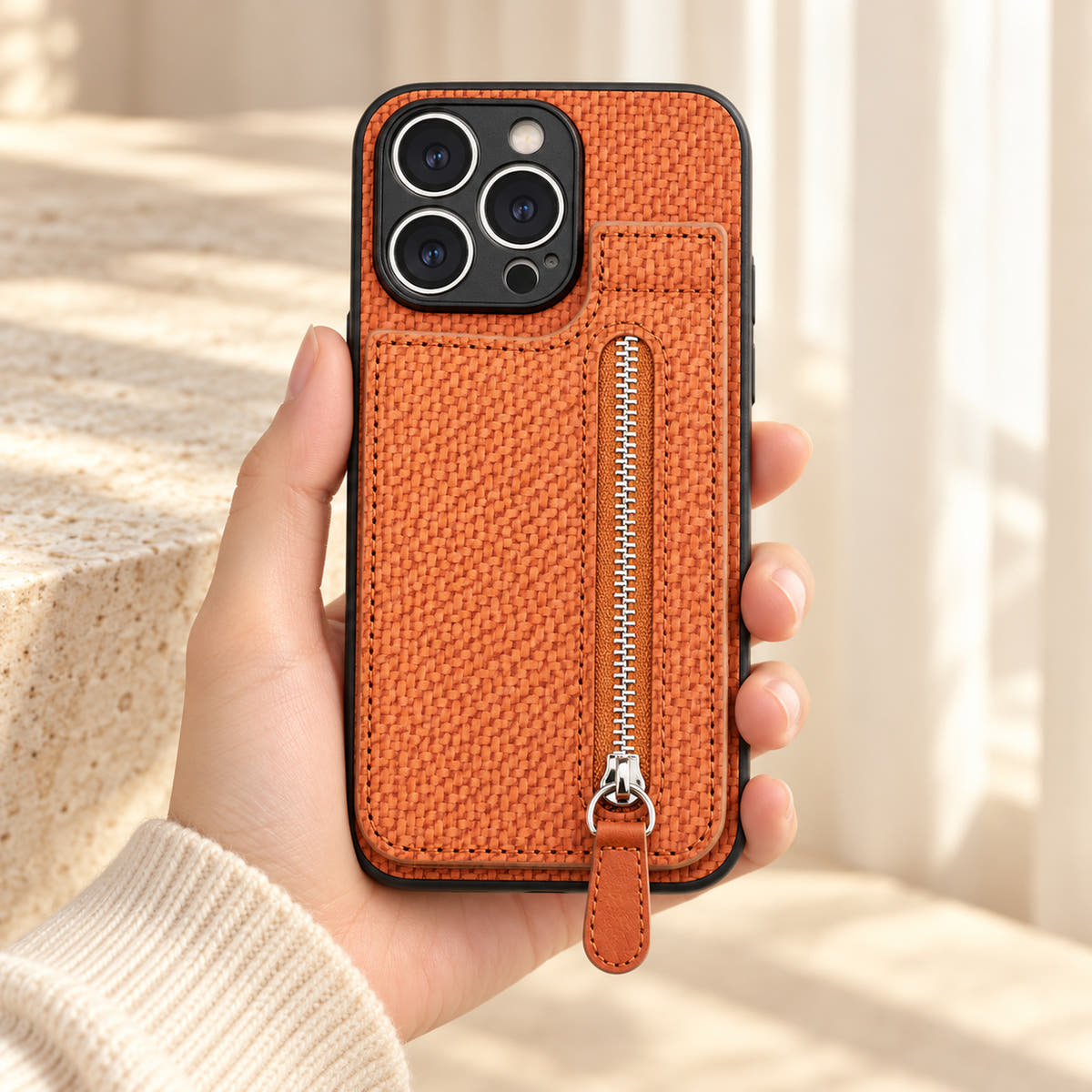 Signature Brown iPhone wallet case held in a cream-sleeved hand, showing its textured rear panel and zip pocket.