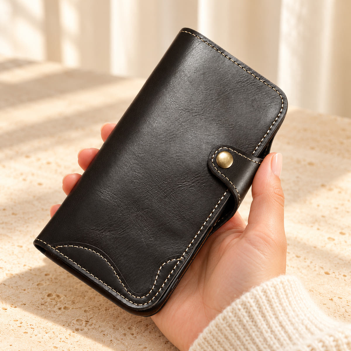 Black stitched iPhone wallet case held in one hand, showing the button tab and curved contrast stitching