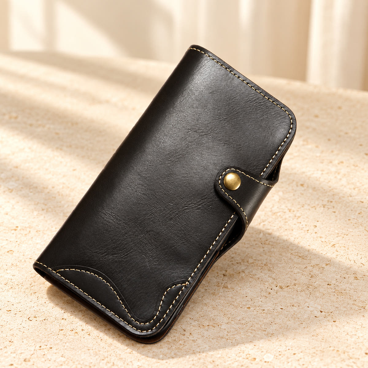 Black stitched iPhone wallet case resting on cream limestone with its front folio and button closure visible